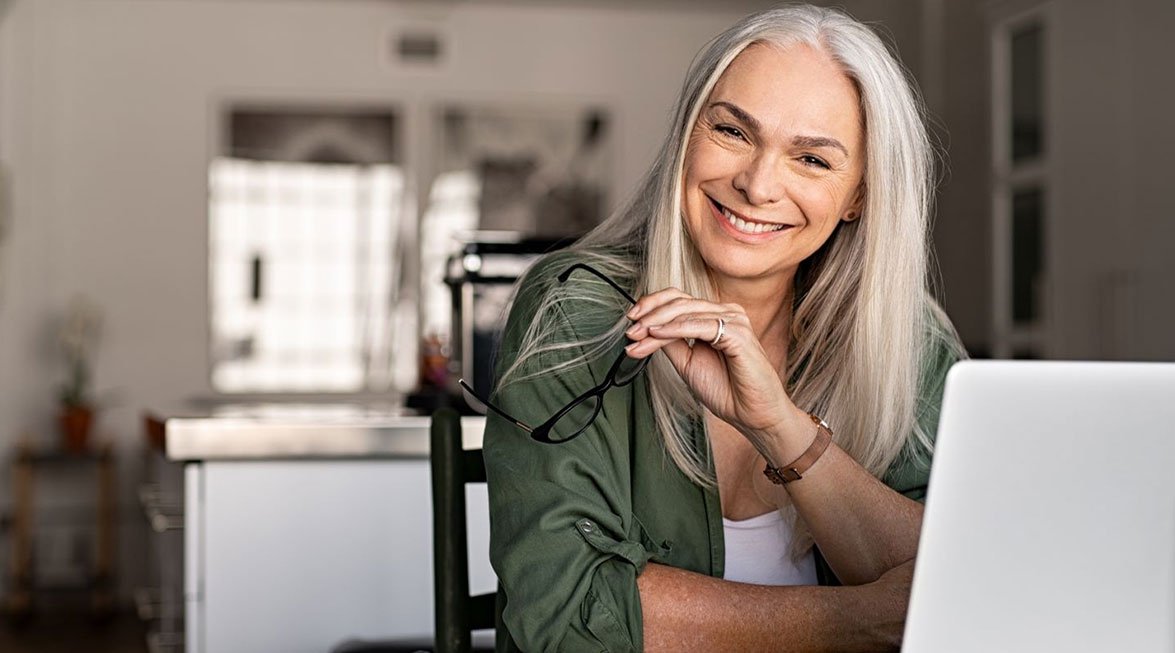  Ältere Dame lacht vor einem Tablet. In der Hand hält sie eine Brille, auf dem Tisch liegen ein Notizbuch und ein Kugelschreiber. Im Hintergrund sind unscharf Fenster und Schränke zu sehen. 