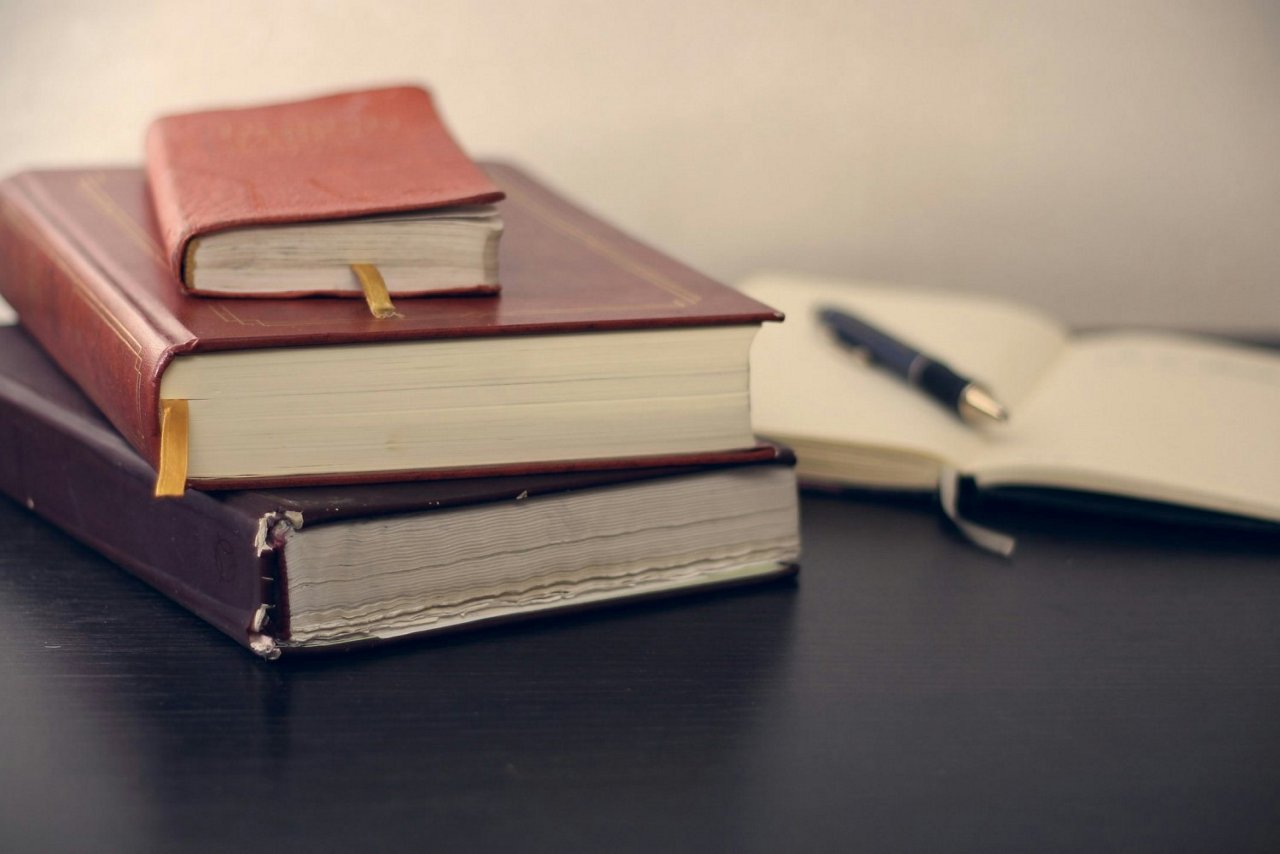 Drei Bücher in verschiedenen Größen, zwei rote und ein braunes, liegen auf einem Tisch. Ein offenes Notizbuch und ein Stift sind daneben.