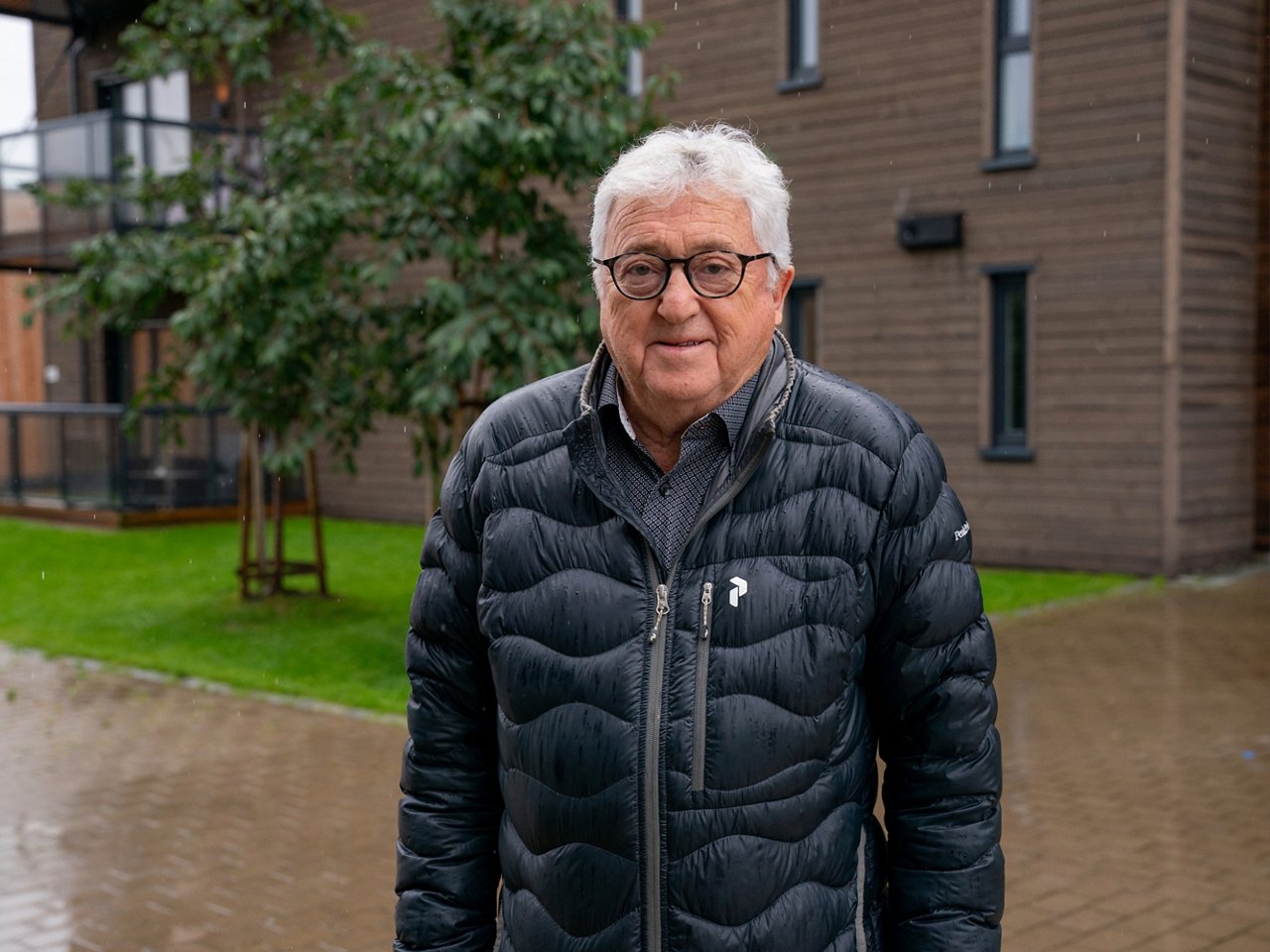 Mann med briller og sort jakke står å smiler foran en bygning.