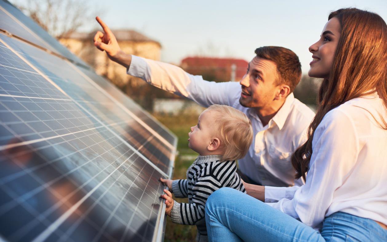 Junge Familie sitzt vor einem Dach mit Solarpanels und lacht.
