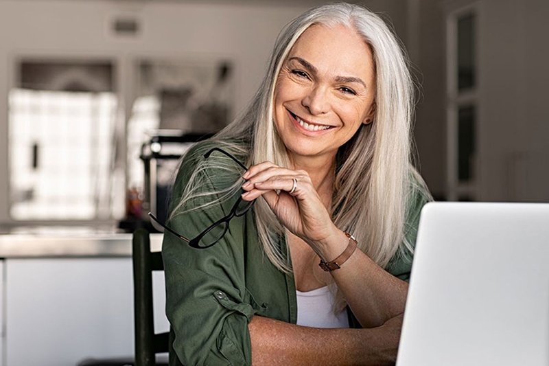 Une femme âgée aux longs cheveux gris est assise en souriant devant son ordinateur portable, elle tient des lunettes à la main - un carnet de notes et un stylo sont posés sur la table.