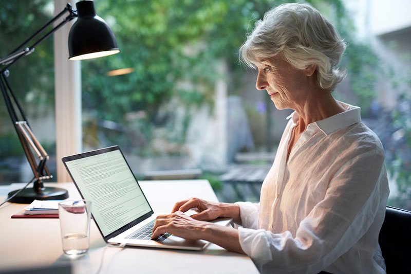 Ältere Dame sitzt am Laptop neben ihr steht ein Wasserglas und eine Tischlampe.