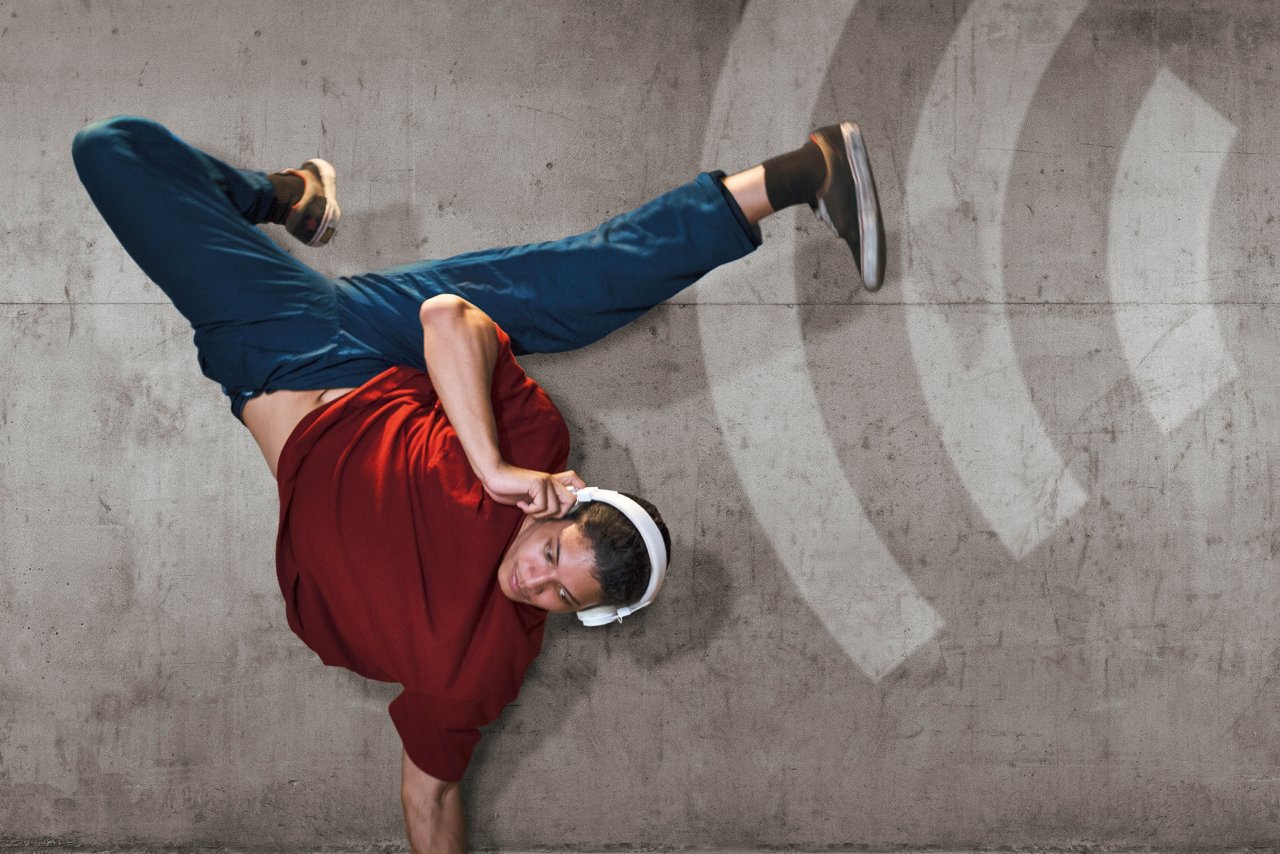 Ein junger Mann tanzt Breakdance vor einer Betonwand mit Funkwellen. 