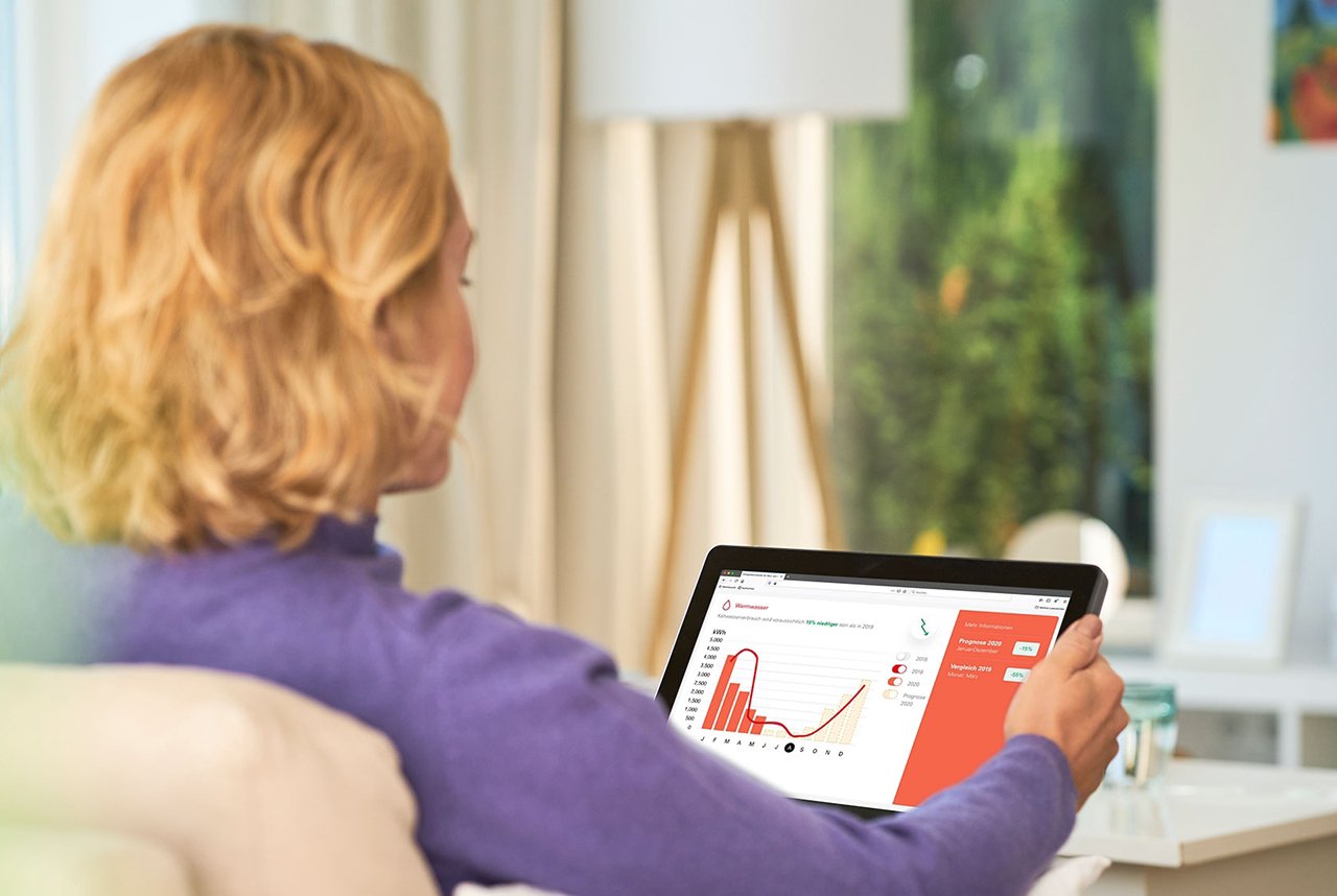Persona usando una tableta en un sofá. La pantalla de la tableta muestra gráficos de barras y líneas, junto con un panel rojo que parece ser parte de una aplicación de análisis de datos. El entorno es doméstico, con una ventana grande al fondo que permite ver vegetación exterior.