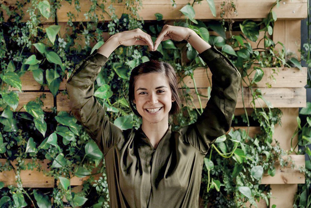 En dame står foran en frodig grønn plantevegg. Hun har løftet hendene over hodet og formet dem til et hjerte. 