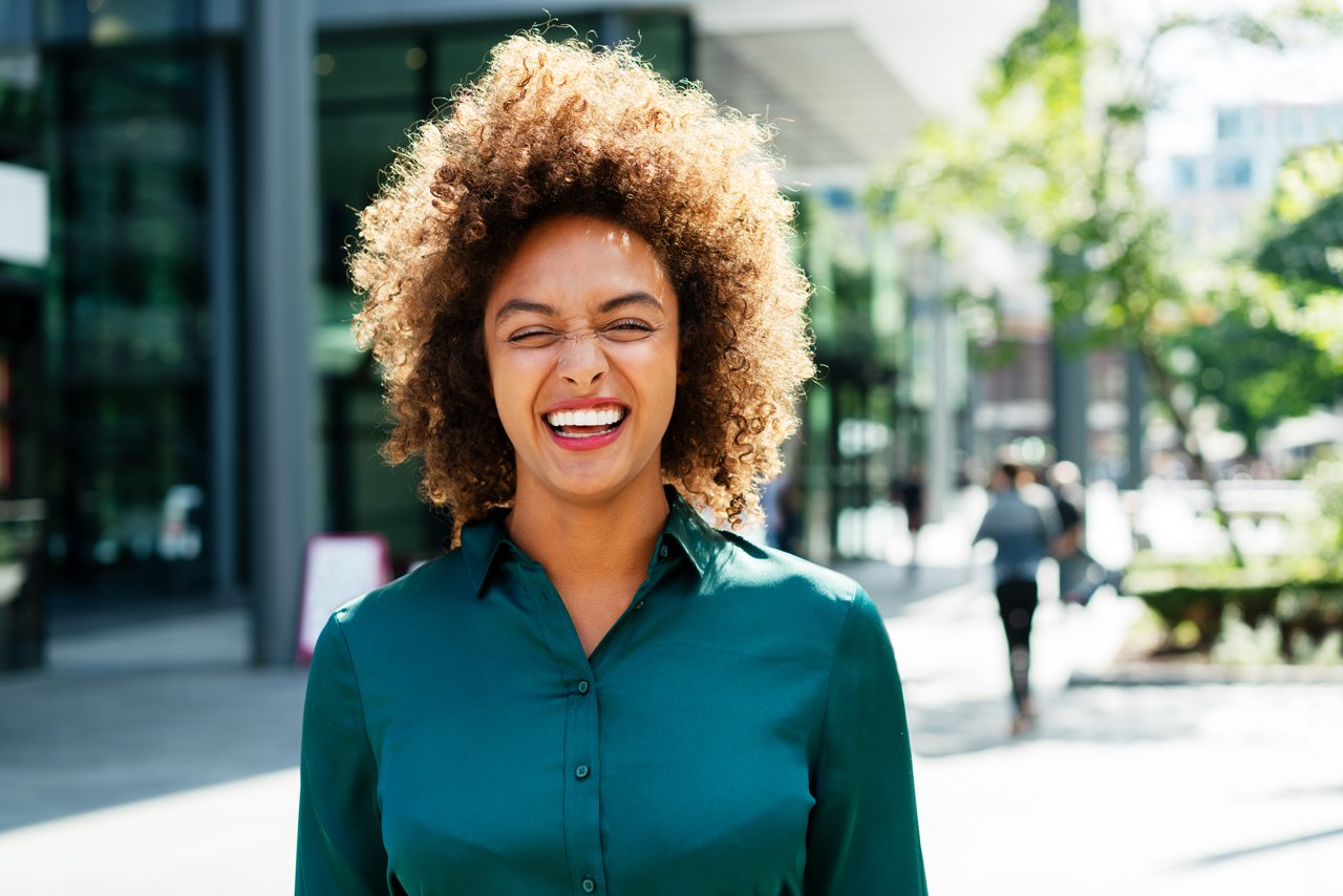 En dame står foran en bygning, hun har et stort smil og myser øynene.