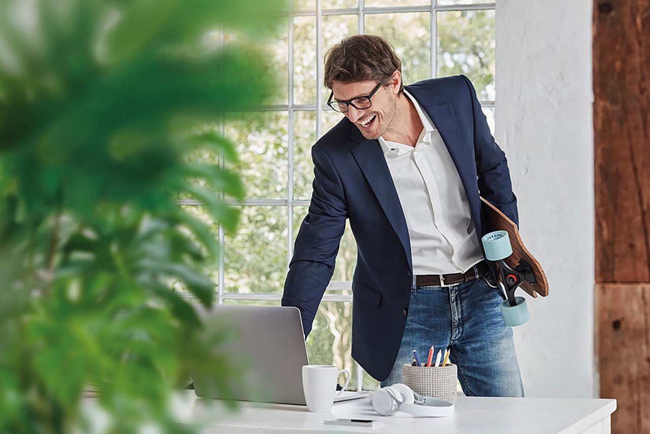 Lachender Mitarbeiter stehend vor Laptop mit einem Getränk in der Hand.