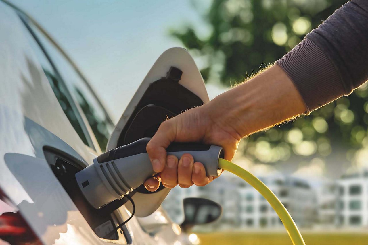 Links im Bild steht ein Auto, schräg von hinten zu sehen. Ein Arm reicht von rechts ins Bild und hält den Ladeanschluss am Elektroauto. Im Hintergrund sind ein weisses Haus, ein grüner Baum und blauer Himmel zu sehen.