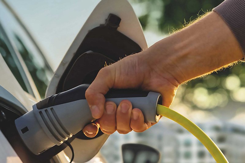 Links im Bild steht ein Auto, schräg von hinten zu sehen. Ein Arm reicht von rechts ins Bild und hält den Ladeanschluss am Elektroauto. Im Hintergrund sind ein weisses Haus, ein grüner Baum und blauer Himmel zu sehen.