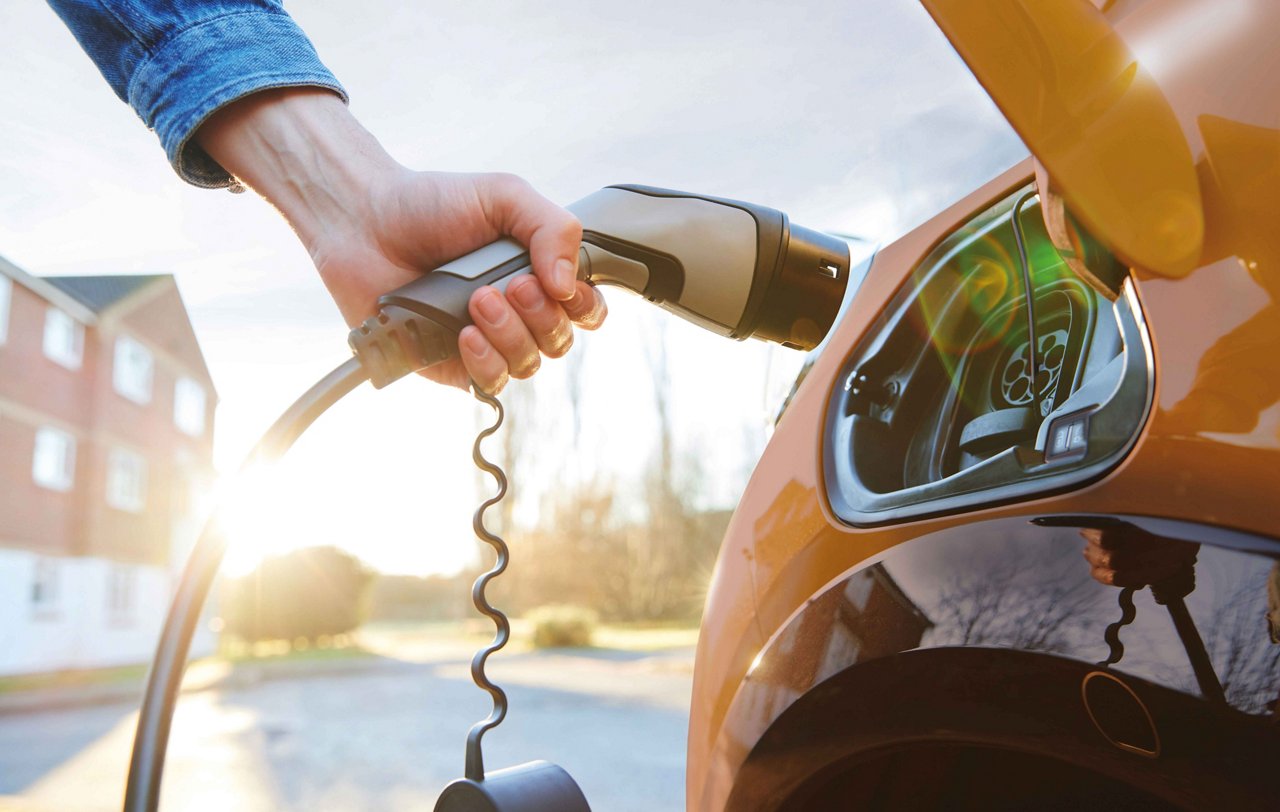 Rechts im Bild steht ein gelbes Elektroauto. Ein Arm hält den Ladestecker und führt ihn zum Ladeanschluss. Im Hintergrund sind ein Haus, einige Bäume und Sonnenschein zu sehen.