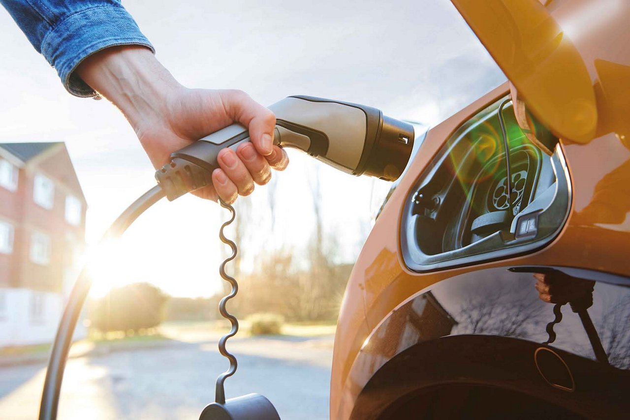 Rechts im Bild steht ein gelbes Elektroauto. Ein Arm hält den Ladestecker und führt ihn zum Ladeanschluss. Im Hintergrund sind ein Haus, einige Bäume und Sonnenschein zu sehen.