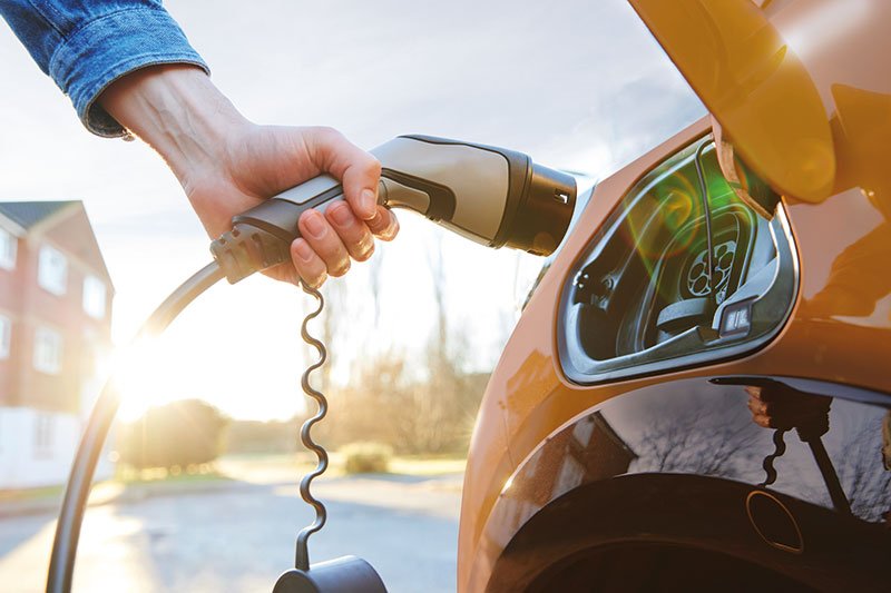 Rechts im Bild steht ein gelbes Elektroauto. Ein Arm hält den Ladestecker und führt ihn zum Ladeanschluss. Im Hintergrund sind ein Haus, einige Bäume und Sonnenschein zu sehen.