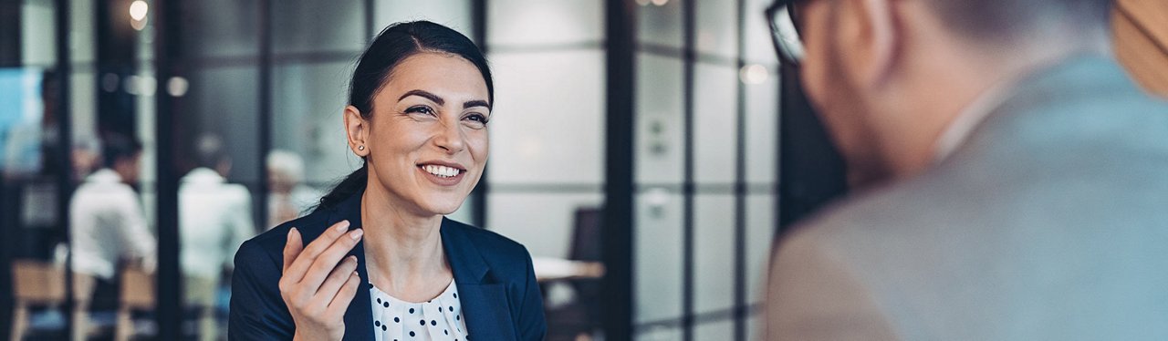 En dame som smiler varmt mens hun engasjert bruker hendene til å kommunisere. Hun er kledd i en elegant dress, som gir henne et profesjonelt og sofistikert utseende.