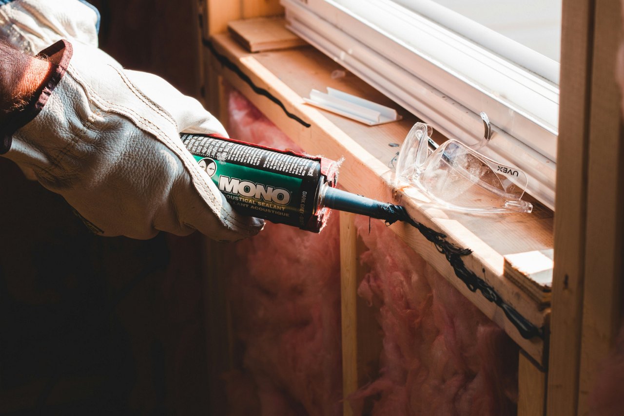  Eine Person mit Arbeitshandschuhen trägt mit einer Kartuschenpistole Dichtmasse an einem Fensterrahmen auf.  Das Bild zeigt eine Baustelle mit Dämmmaterial und Holzverkleidung im Hintergrund.