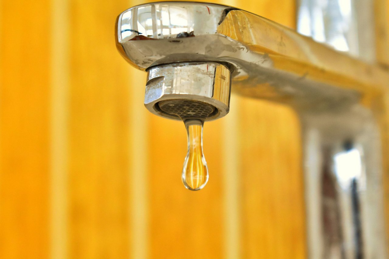 Ein Wassertropfen hängt am metallischen Wasserhahn vor gelb-orangem, unscharfem Hintergrund.