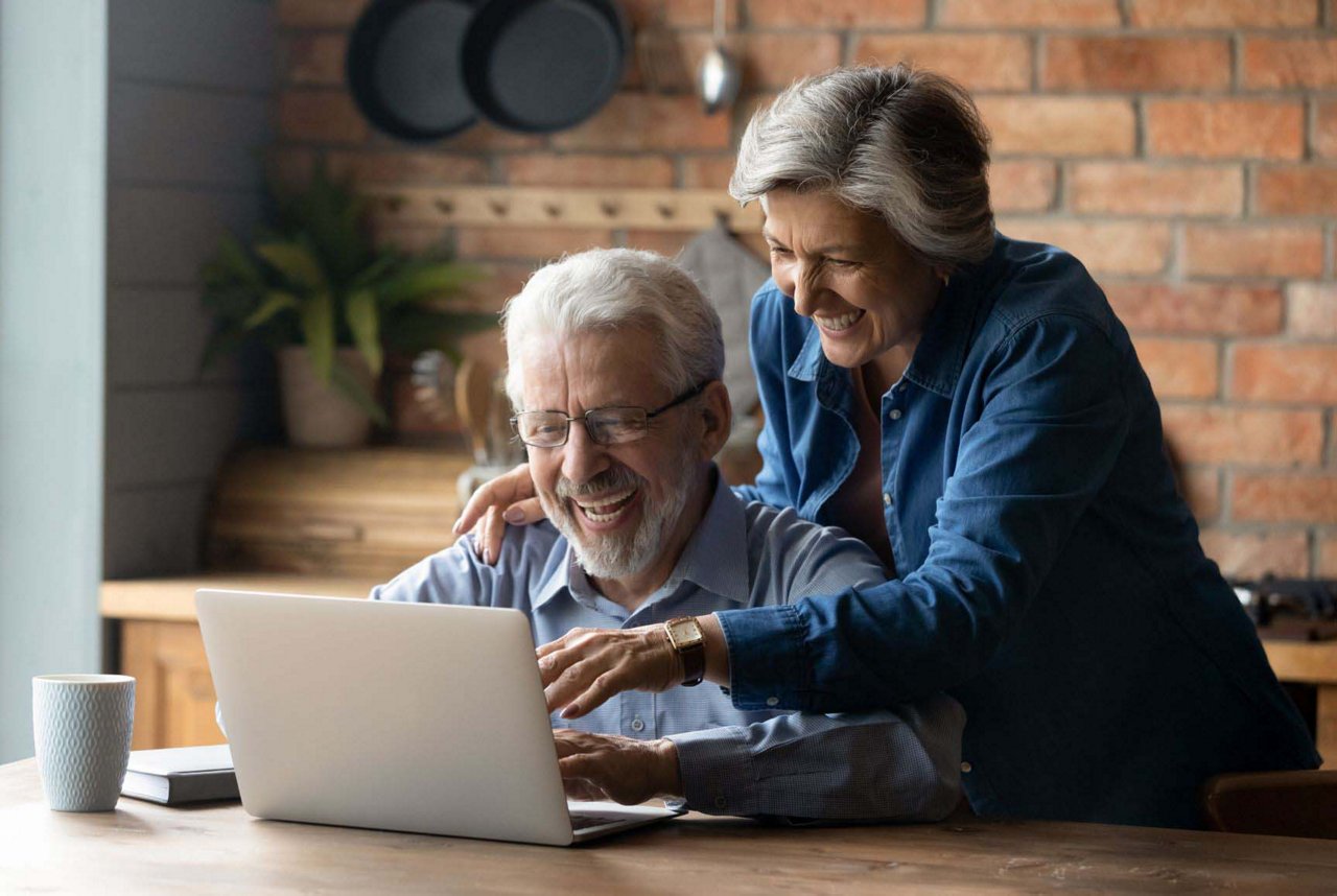 Un couple âgé se tient dans la cuisine. L'homme cuisine et regarde avec intérêt l'ordinateur portable que manipule sa femme. Tous deux sourient et semblent satisfaits.