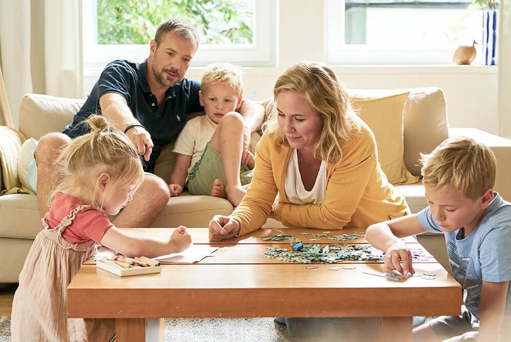 Eltern und drei Kinder sitzen im Wohnzimmer. Die Mutter und der Sohn puzzeln, die Tochter malt, während der Vater mit dem jüngeren Sohn zusieht.