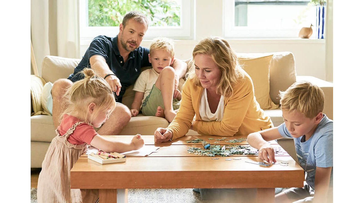 Eltern und drei Kinder sitzen im Wohnzimmer. Die Mutter und der Sohn puzzeln, die Tochter malt, während der Vater mit dem jüngeren Sohn zusieht.
