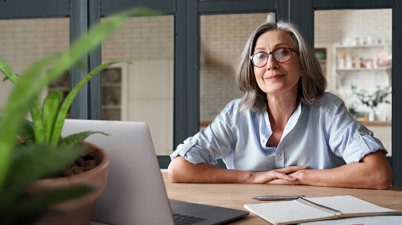  Frau mit grauem Haar und Brille sitzt entspannt an einem Schreibtisch. Vor ihr liegen ein Laptop, ein Notizbuch und ein Stift.