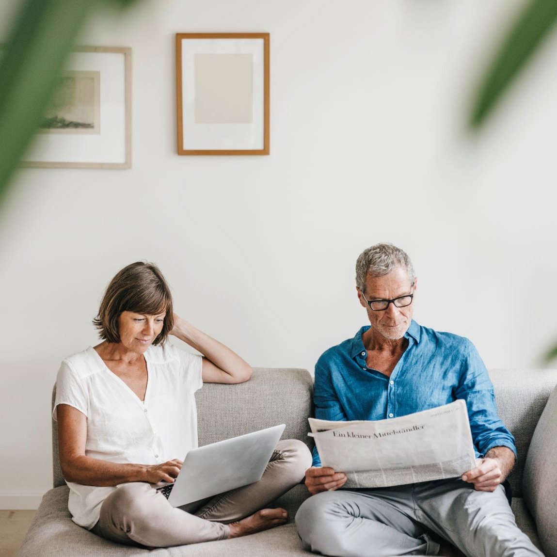 Un couple âgé est assis sur un canapé gris. Elle est sur son ordinateur portable, il lit le journal. 