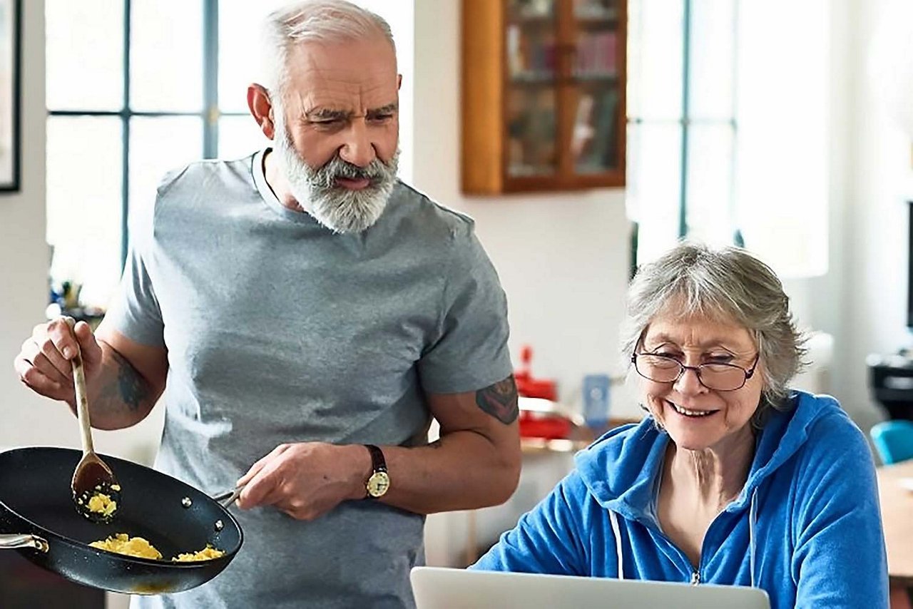 Ein älteres Paar in der Küche: Die Frau lächelt am Laptop, während der Mann Rührei zubereitet. Die helle Umgebung strahlt eine warme, harmonische Alltagsstimmung aus.