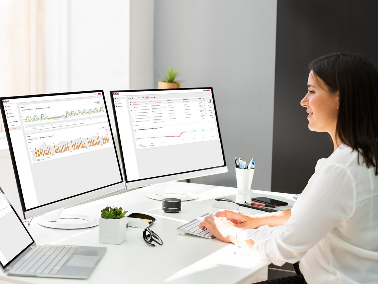 Frau sitzt am Schreibtisch und arbeitet an zwei Monitoren mit Diagrammen und Datenanalysen in einem modernen Büro