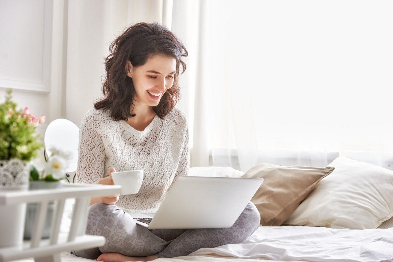 Eine Frau mit braunen schulterlangen Haaren sitzt mit Laptop auf dem Schoss und einer Tasse in der Hand auf Bett. Sie lächelt in ihr Laptop.