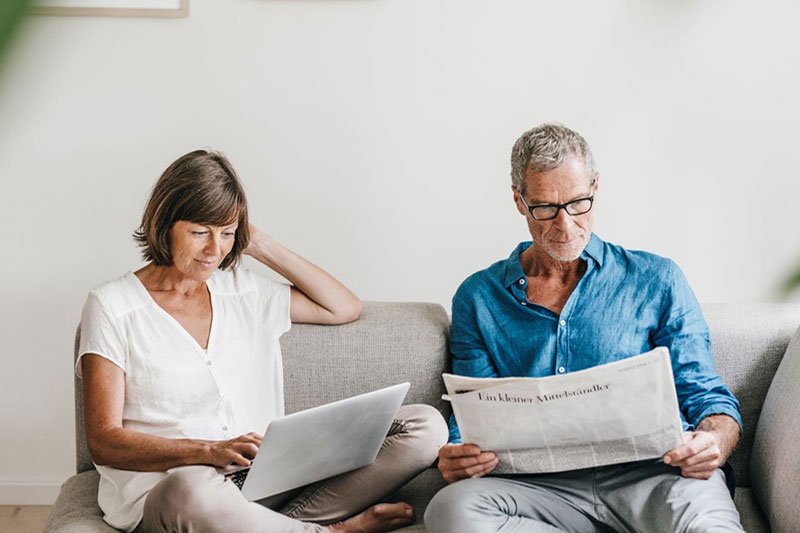 Ein älteres Paar sitzt auf einem grauen Sofa. Sie ist am Laptop, er liest Zeitung. 