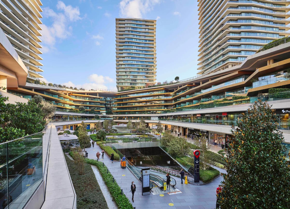 Mehrstöckige Gebäude mit Balkonen und großzügigen Glasflächen umrahmen einen Innenhof mit viel Grün, Gehwegen und einigen wenigen Personen