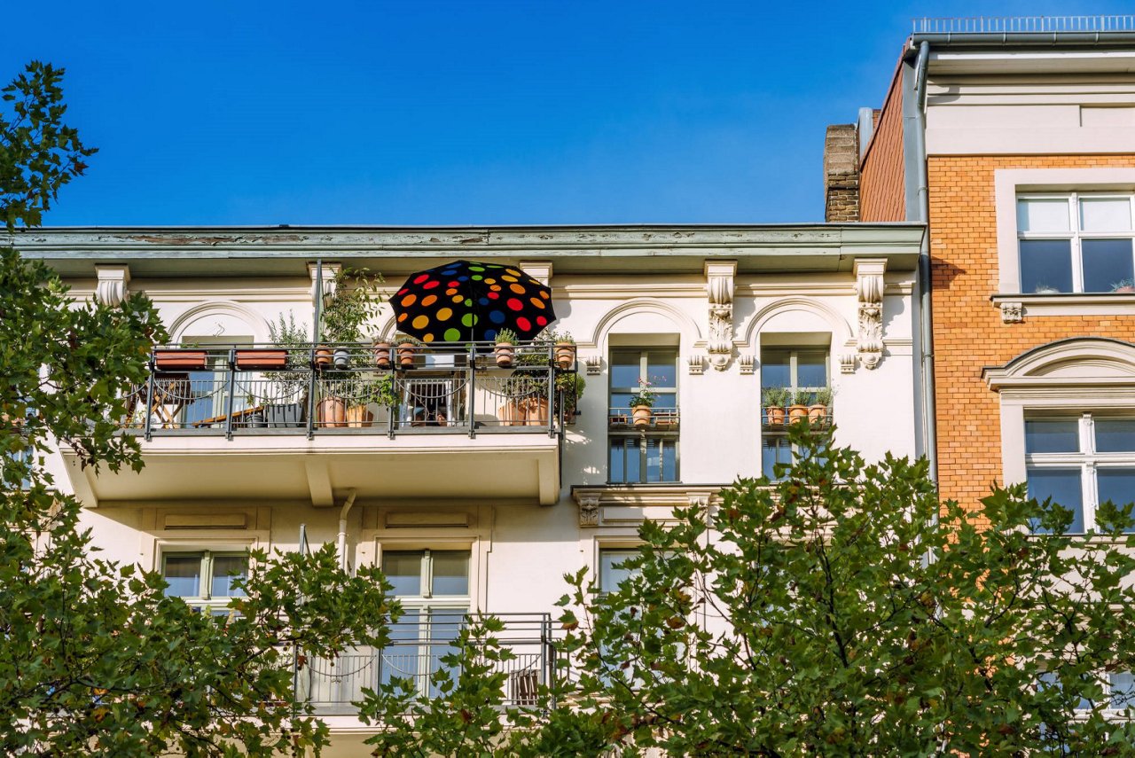 Kleines Mehrfamilienhaus mit Balkon und buntem Regenschirm in einer Häuserzeile, umgeben von grünen Bäumen