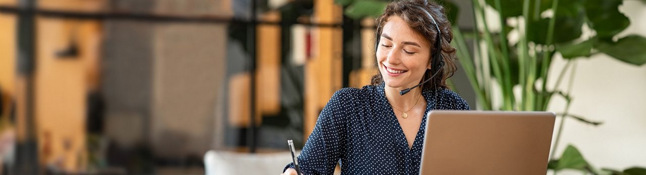 Lächelnde Mitarbeiterin mit Headset am Laptop im Kundenservice, macht sich Notizen während eines Gesprächs
