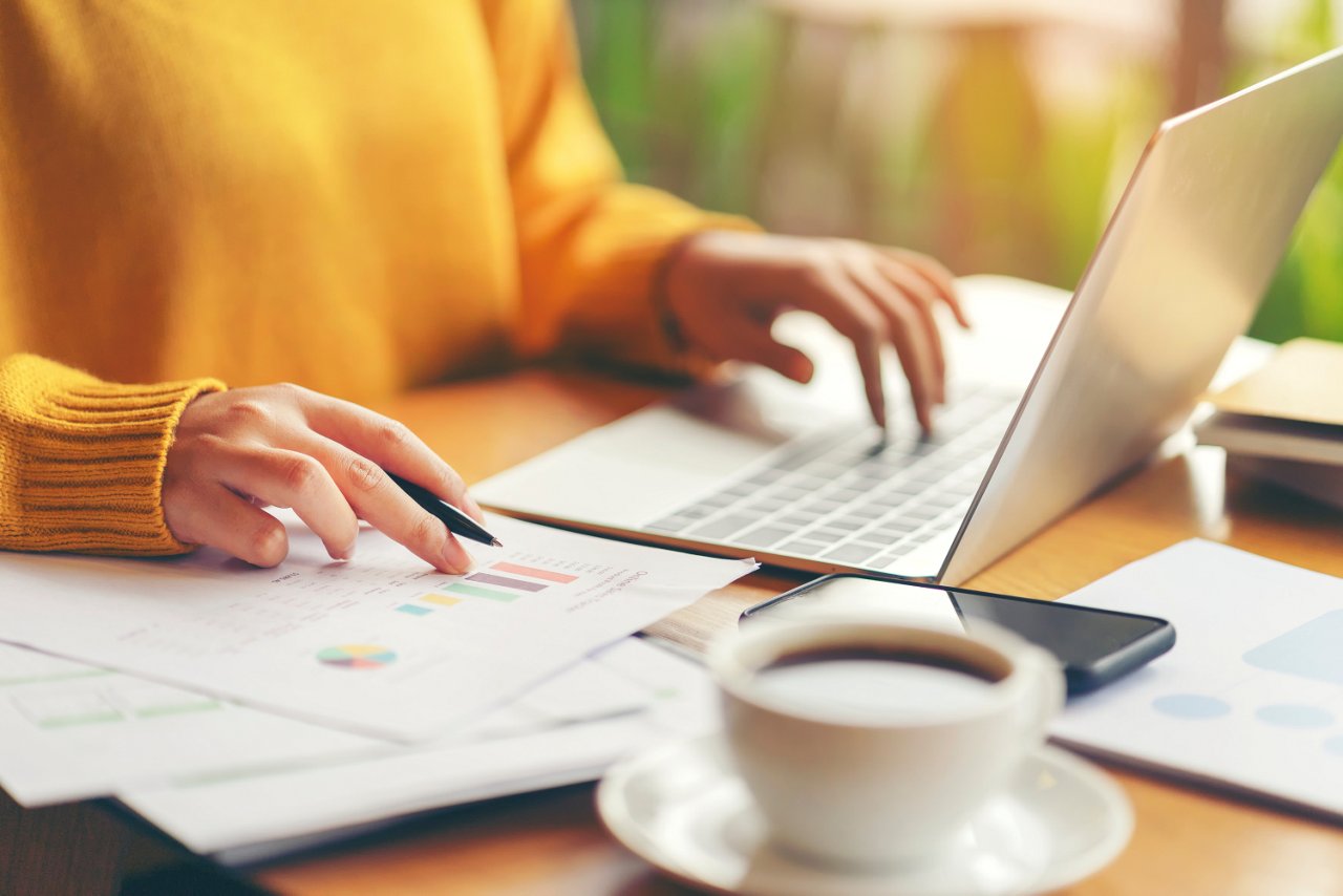 Eine Person in einem gelben Pullover tippt auf einem Laptop und überprüft ein Diagramm mit einem Stift. Auf dem Tisch liegen Unterlagen, ein Smartphone und eine Kaffeetasse.