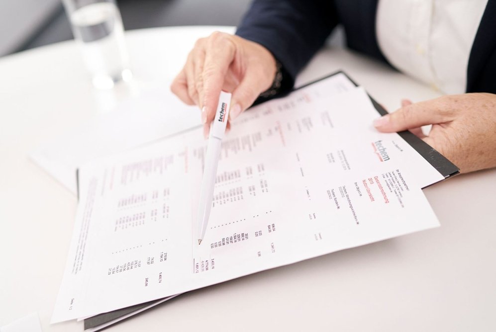 Manos de una persona  sosteniendo y señalando una liquidación de Techem con varias páginas que contiene tablas y texto, con información sobre el consumo de calefacción. La persona está usando un bolígrafo para señalar el documento.