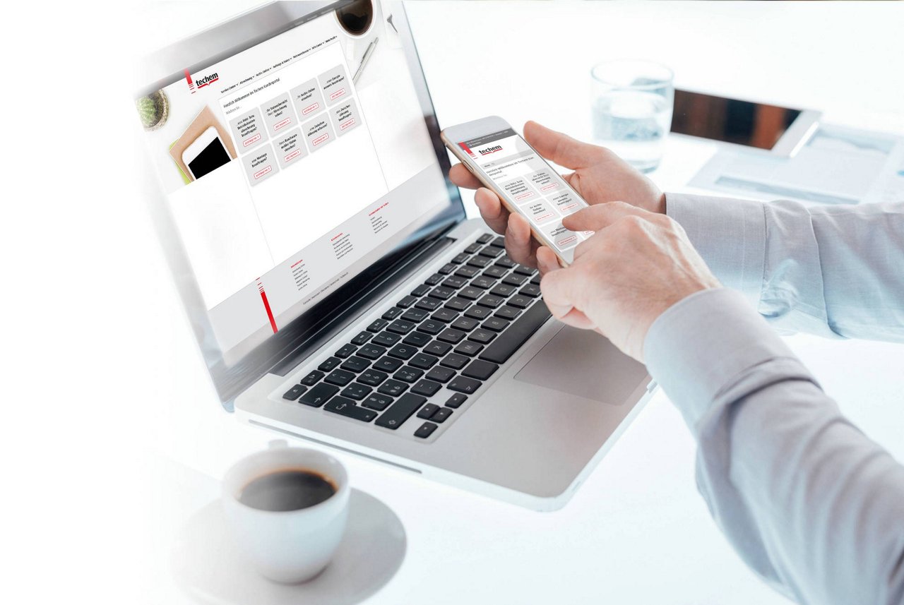 Person hält Smartphone mit Kalender-App vor Laptop mit geöffnetem Kalender, Kaffeetasse steht daneben