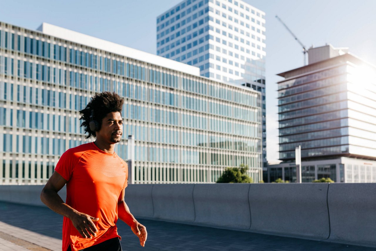 Jungen Mann mit Afro-Frisur joggt in einem orangen T-Shirt und Kopfhörern durch eine moderne Stadtlandschaft.