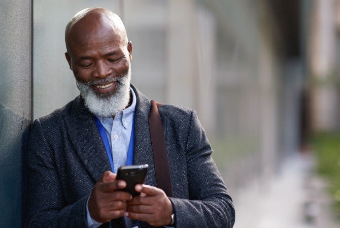 Mann lächelt, lehnt an einer Hauswand und hält sein Handy in der Hand
