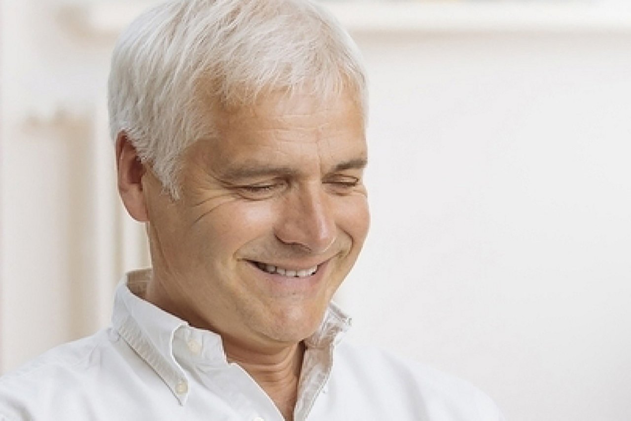 Un homme âgé en chemise blanche est assis devant son ordinateur portable en riant.