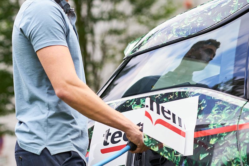 Ein Mann lädt ein Techem-Elektroauto. Er trägt ein blaues T-Shirt. Im Hintergrund steht ein Baum.