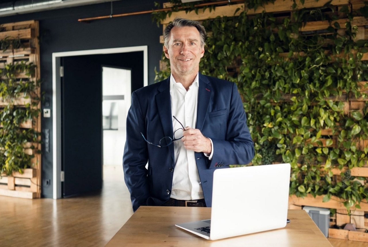 Mann mit blauem Jackett steht vor einer grünen Wand, hält seine Brille in der Hand. Auf dem Tisch steht ein Laptop