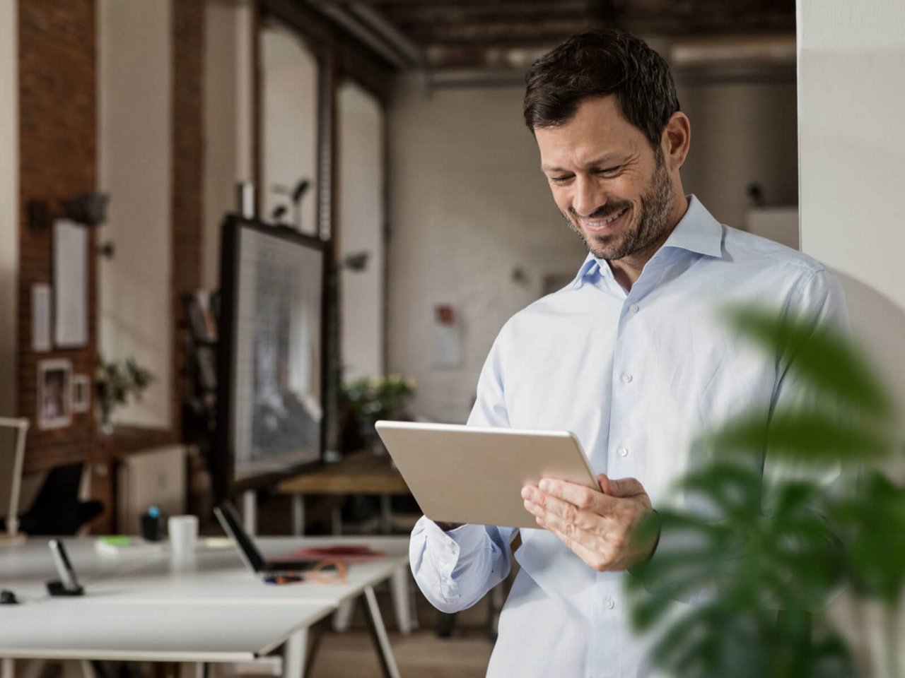 Mann in weißem Hemd lächelt und nutzt Tablet in modernem Büro mit Schreibtischen und Pflanzen