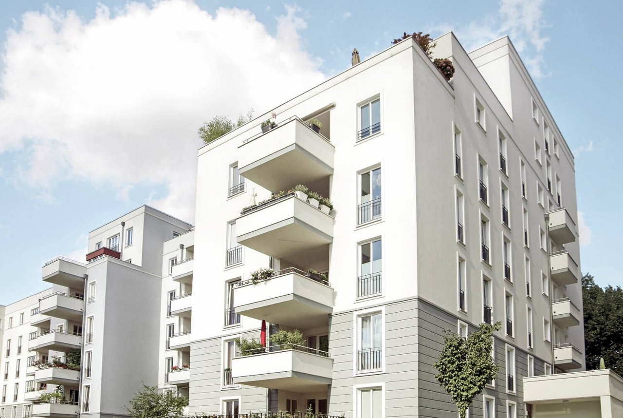 Ein modernes, mehrstöckiges Wohngebäude mit weißen Fassaden und mehreren Balkonen, die mit Pflanzen dekoriert sind.