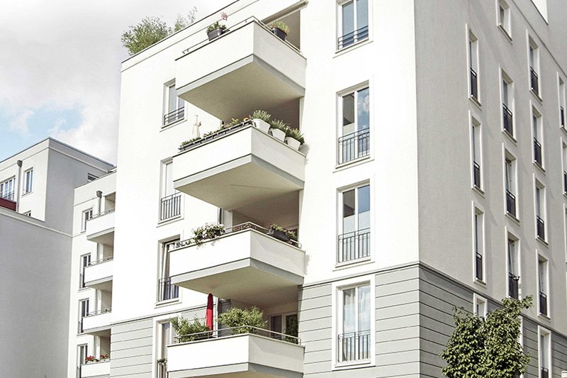Modernes, mehrstöckiges Mehrfamilienhaus mit Balkonen, weissen Fassaden und grünen Pflanzen vor blauem Himmel.