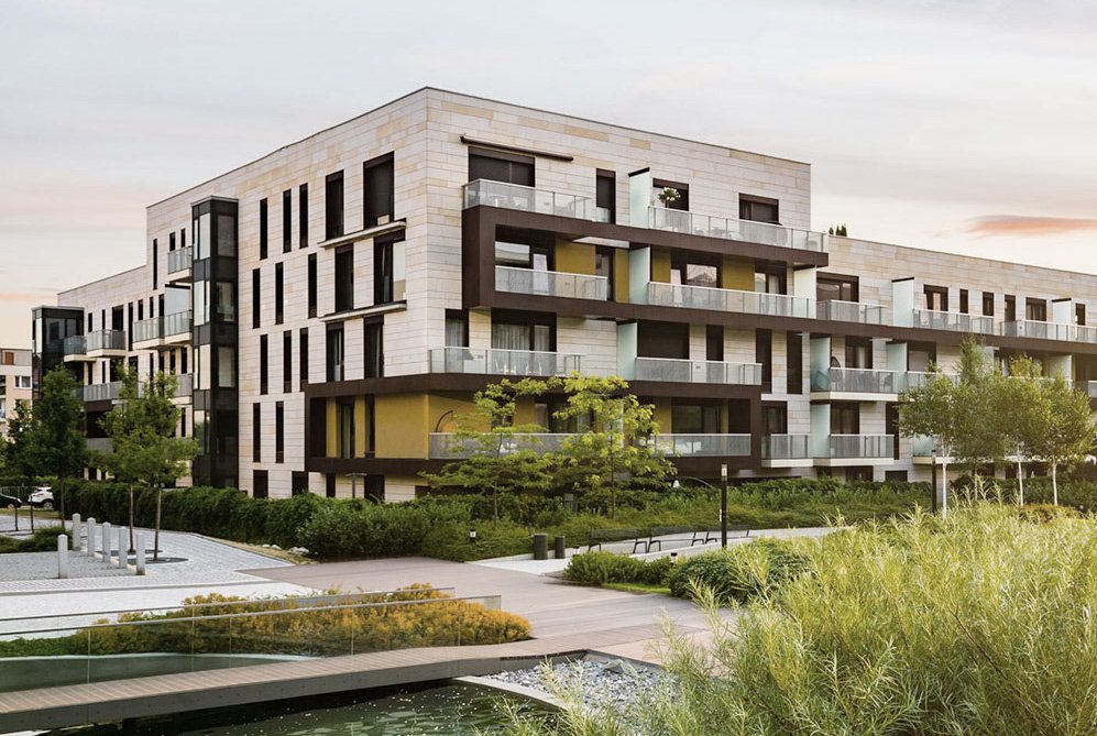 Wohngebäude mit großen Glasbalkonen und heller Fassade. Davor ein angelegter Park mit Bäumen, Wegen und einem Wasserlauf.