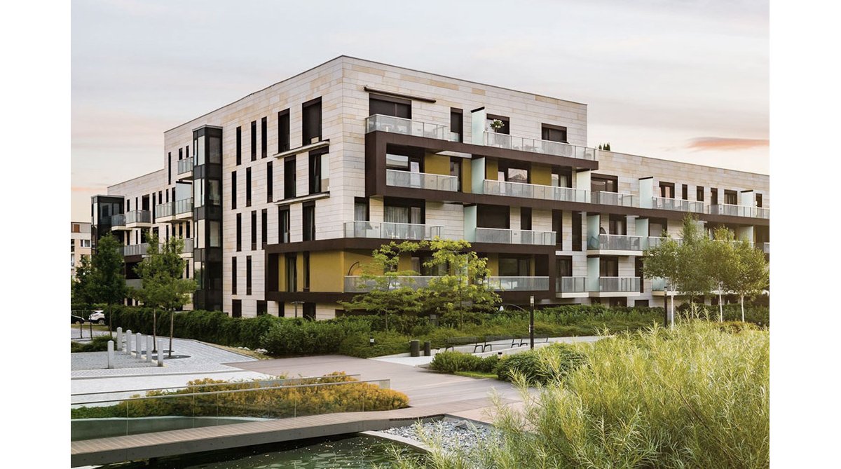 Wohngebäude mit großen Glasbalkonen und heller Fassade. Davor ein angelegter Park mit Bäumen, Wegen und einem Wasserlauf.