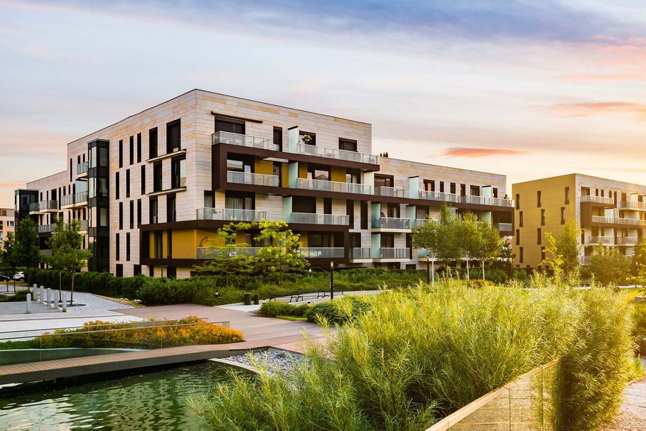 Eine idyllische Wohnsiedlung bei Sonnenuntergang. Zu sehen sind Teiche, Sitzbänke und viel Begrünung.