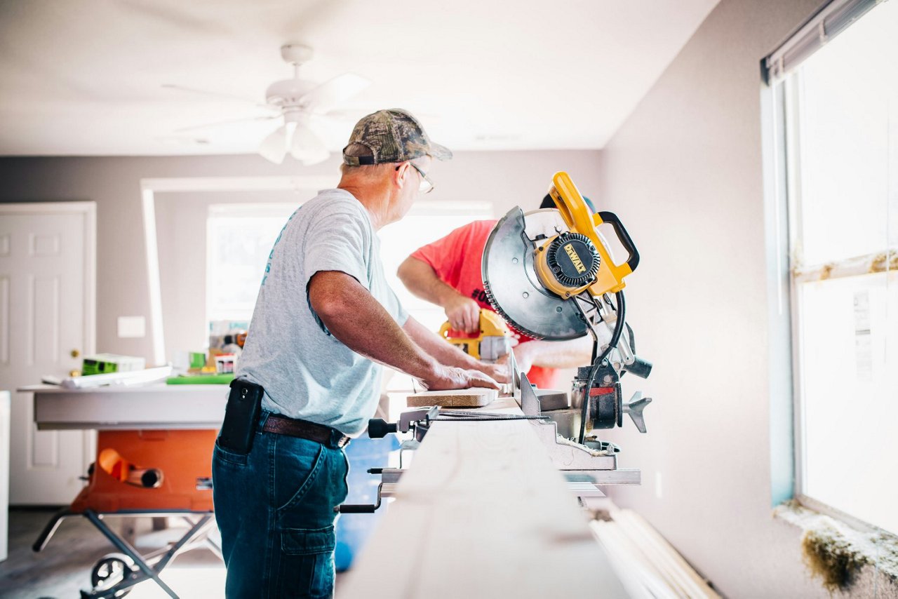 Zwei Handwerker schneiden Holz in einem renovierungsbedürftigen Raum. Einer bedient eine Kappsäge, der andere arbeitet mit einem Elektrowerkzeug. 