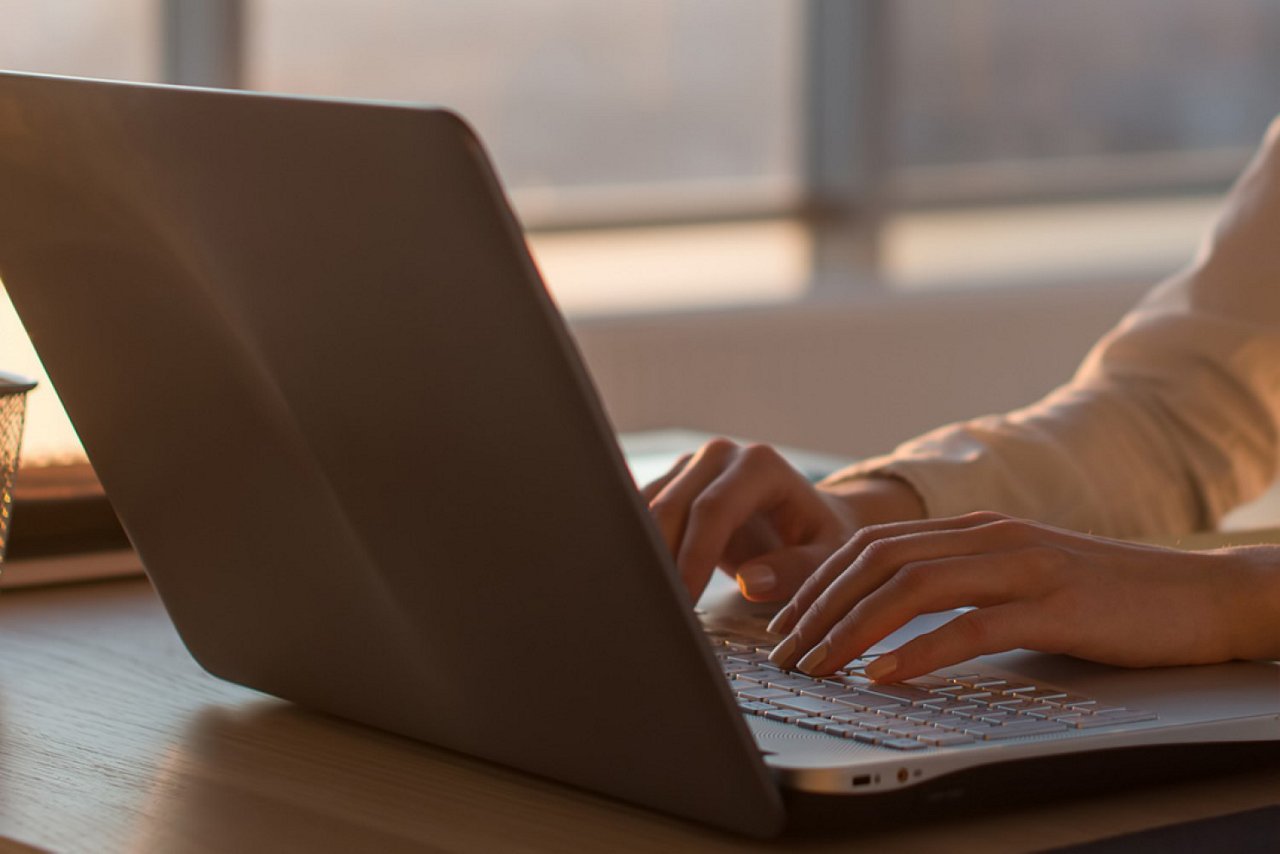 Die Hände der Person sind auf der Tastatur zu sehen, während der Laptop auf einem Holztisch steht.