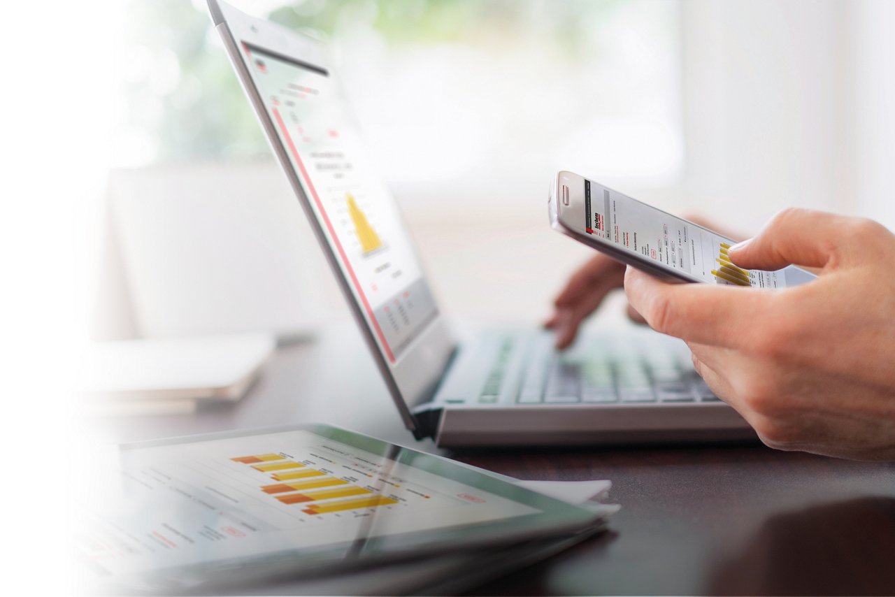 Persona usando un smartphone frente a una computadora portátil abierta, ambos mostrando gráficos y diagramas. También hay una tablet sobre la mesa con datos gráficos similares. El fondo está desenfocado para centrar la atención en los dispositivos y las manos de la persona