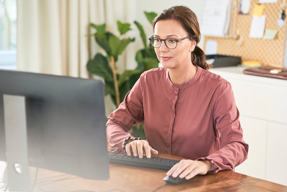 Mujer adulta con gafas trabajando en un ordenador de escritorio en casa. Escritorio de madera con teclado, ratón y monitor.