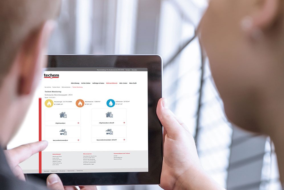 Dos personas sosteniendo y mirando una tablet. La pantalla de la tablet muestra una página web de "techem" con varias seccionese íconos que representan diferentes servicios como calefacción, agua y gestión de energía.